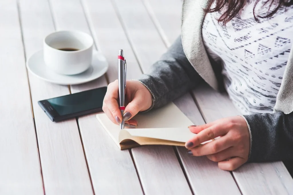 Woman in her 50s writing questions in a notebook at a sunlit kitchen table